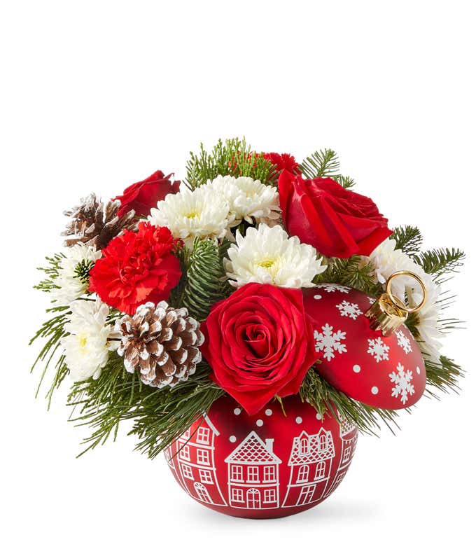 Holiday floral arrangement with red roses, red carnations, white poms, seasonal greenery, and pinecones. Displayed in a red ornament-shaped container with white village and snowflake details.