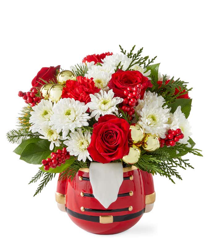 Festive floral arrangement with red roses, miniature red carnations, white poms, seasonal greenery, red berry picks, and gold ornaments in a red Nutcracker-themed container.