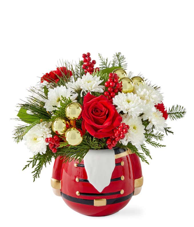 Festive floral arrangement with red roses, miniature red carnations, white poms, seasonal greenery, red berry picks, and gold ornaments in a red Nutcracker-themed container.