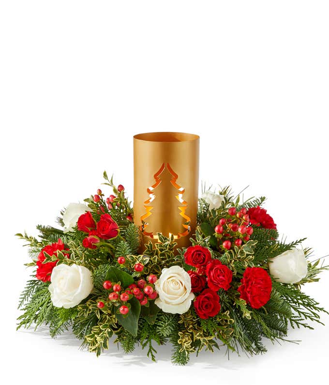 Holiday centerpiece arrangement featuring white roses, miniature red carnations, red hypericum berries, and seasonal greenery surrounding a gold metal candle holder with a cut-out tree design.