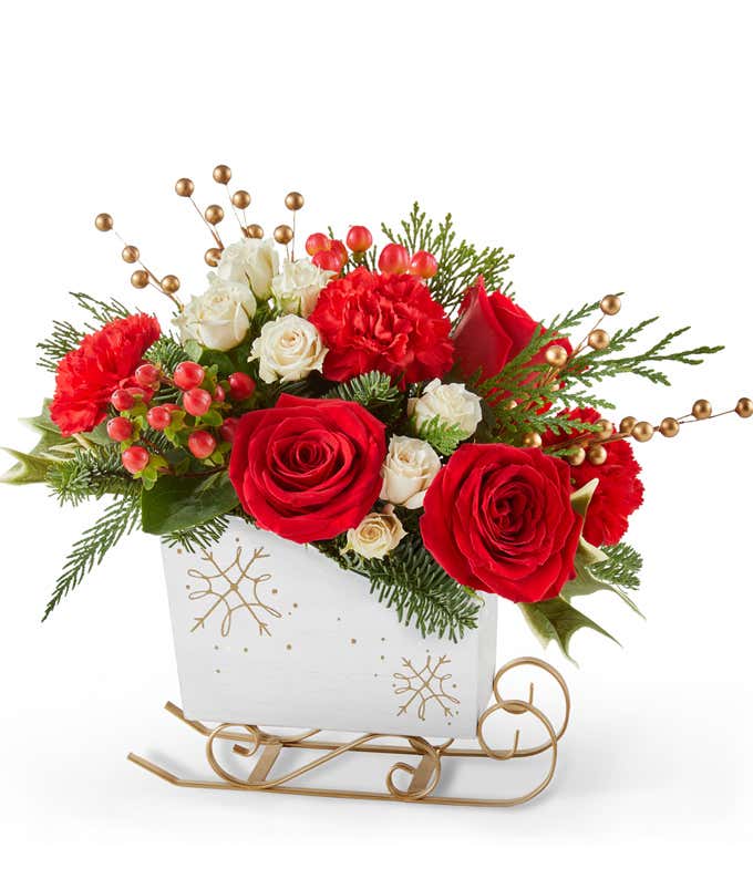 Festive holiday arrangement with red roses, red carnations, white spray roses, and red hypericum berries, accented by gold berry picks and seasonal greenery in a white sleigh with gold details.