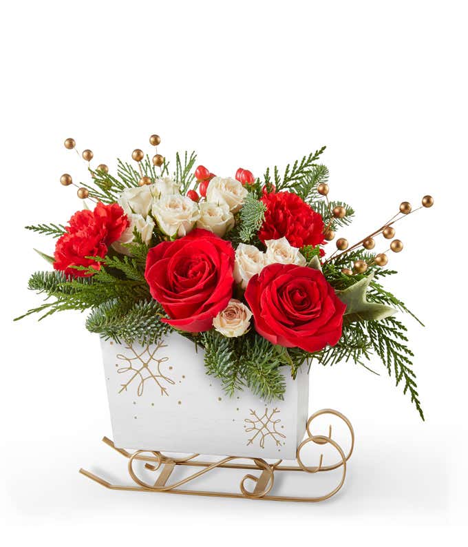 Festive holiday arrangement with red roses, red carnations, white spray roses, and red hypericum berries, accented by gold berry picks and seasonal greenery in a white sleigh with gold details.