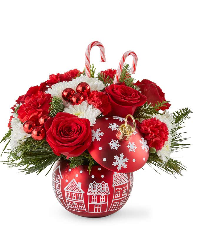 Festive holiday arrangement with red roses, red carnations, white poms, seasonal greenery, and red ornament accents, topped with candy canes. Displayed in a red ornament-shaped container with white holiday village designs.