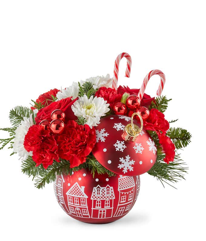 Festive holiday arrangement with red roses, red carnations, white poms, seasonal greenery, and red ornament accents, topped with candy canes. Displayed in a red ornament-shaped container with white holiday village designs.