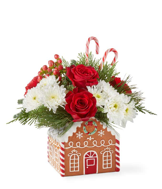 Festive holiday arrangement with red roses, red carnations, white poms, red hypericum berries, and seasonal greenery, accented with candy canes. Displayed in a gingerbread house container with white icing details and candy decorations.