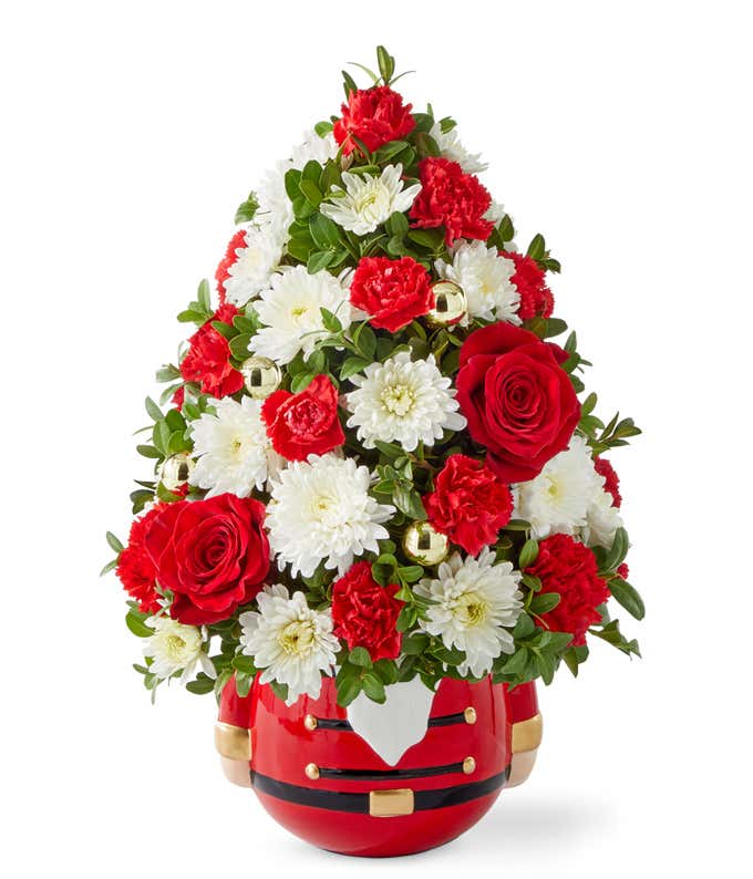 Christmas tree-shaped floral arrangement with red roses, red carnations, white poms, and seasonal greenery, arranged in a festive red Nutcracker-themed container with gold accents.