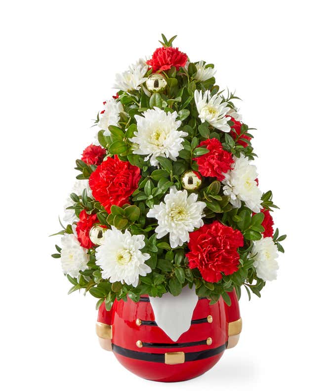 Christmas tree-shaped floral arrangement with red roses, red carnations, white poms, and seasonal greenery, arranged in a festive red Nutcracker-themed container with gold accents.