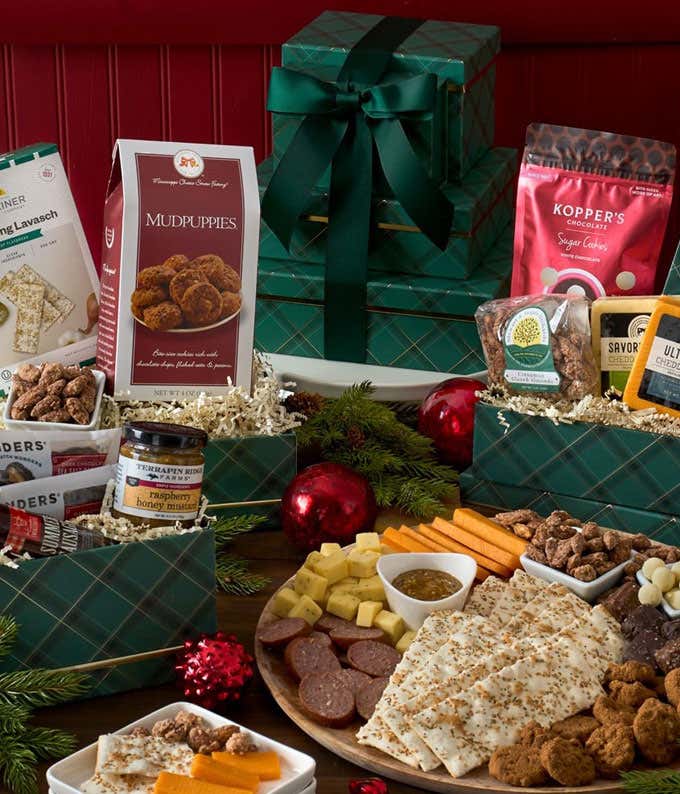 A festive gift tower with green plaid boxes filled with gourmet treats, including cheese, crackers, sausages, chocolate-covered nuts, sugar cookies, mudpuppies, and spreads. Decorated with red ornaments and greenery, perfect for holiday celebrations.