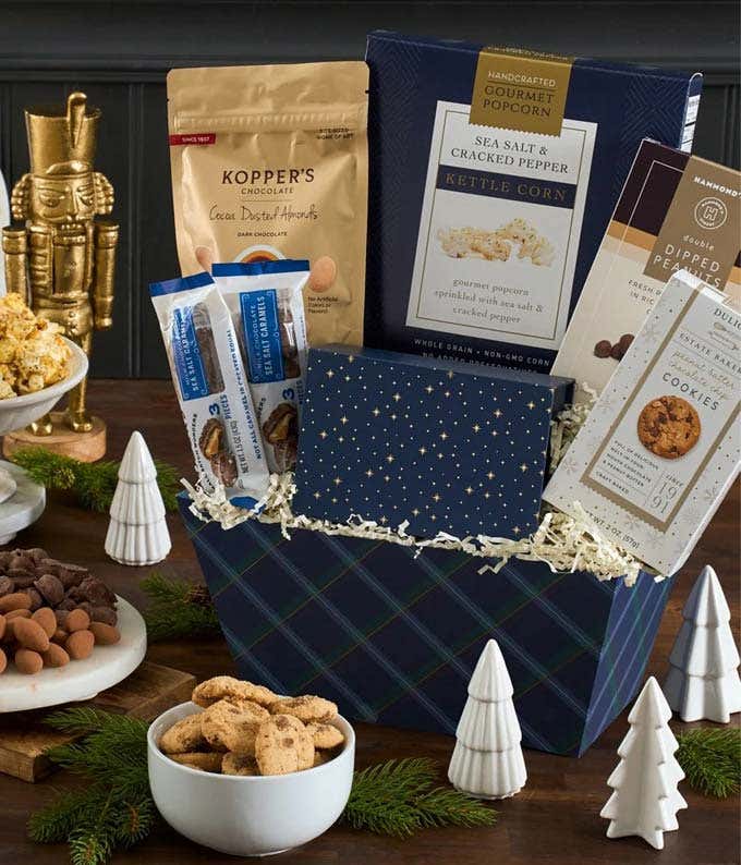 Navy blue basket with black and white ribbon, containing kettle corn, sea salt caramel cookies, Sanders sea salt caramels, Marich dark chocolate sea salt caramels, Rustic Bakery flatbread, smores grahams, assorted cookies, and chocolates. Decorated with b