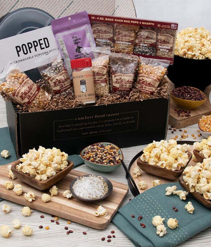 Gift box with 10 bags of popcorn kernels, a microwave popcorn popper, salt & vinegar seasoning, bag of garlic seasoning. In front of the gift basket are 4 small bowls of popped popcorn, 3 small bowls of kernels on a grey wooden table.