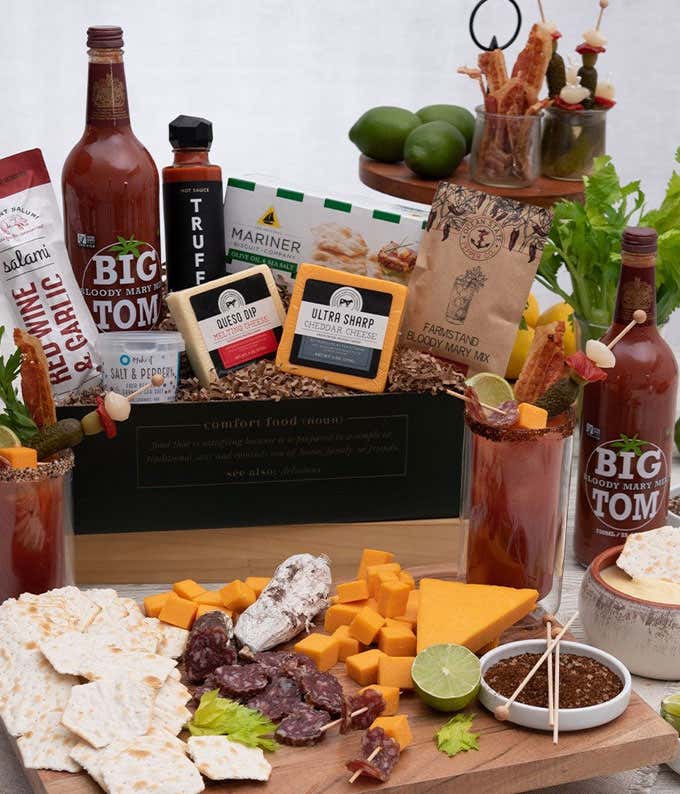 A gift box with blocks of queso & cheddar cheese, packet and bottle of bloody mary mix, salt & pepper seasoning blend, box of water crackers, hot sauce from Truff. In front of the box is a grazing board with crackers, sliced soppressata, cubed cheddar, an
