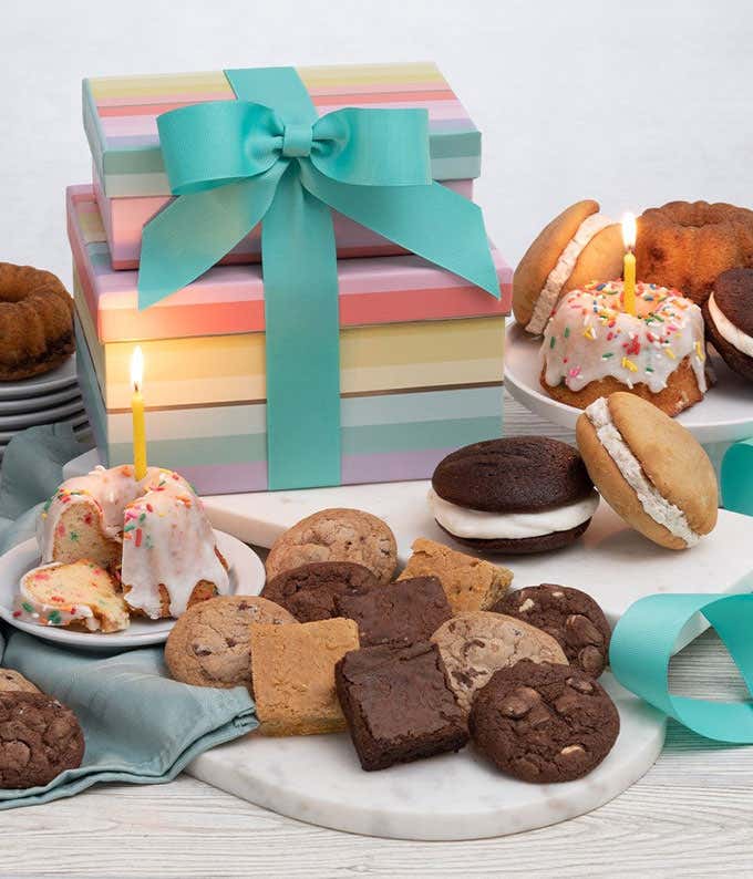 Two rainbow striped gift boxes with a blue bow. In front, a plate with a mini bundt cake with a candle, another tray with cookies, brownies, and blondies. Behind those, a cake tray with whoopie pies and mini bundt cakes. 