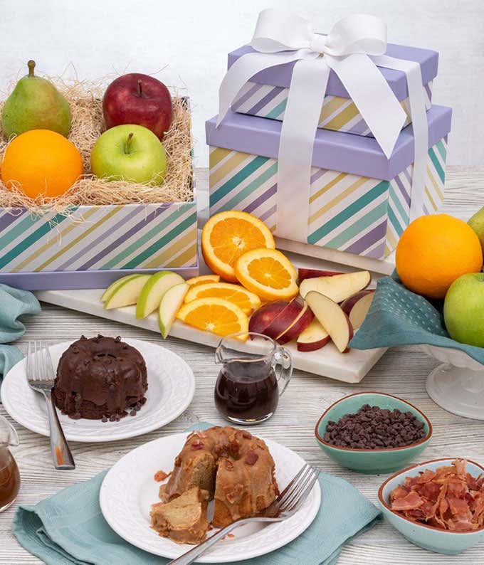 Two pastel striped boxes stacked on top of one another with a white bow. An open box with a pear, red apple, orange, and green apple inside. In front, sliced apples and oranges. In the foreground, on two separate plates, a chocolate mini bundt cake, and a