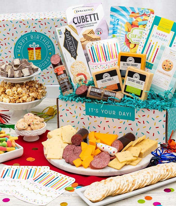 In a gift box, a bag of of chocolate wafter Cubetti's, a box of crackers, a bag of gummy worms, a box of kettle corn, three blocks of cheese, two packages of sausage. In front of the box, a plate of sliced meat & cheese and a plate of crackers. Beside the