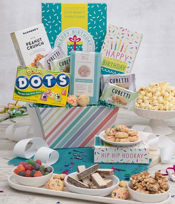 A gift box with a box of kettle corn, peanut crunch, sugar cookies, wafer cookies, and a box of sour DOTS. In front of the gift box, dishes with popcorn, cookies, wafer cookies, DOTS, and peanut crunch lay out on a wooden table