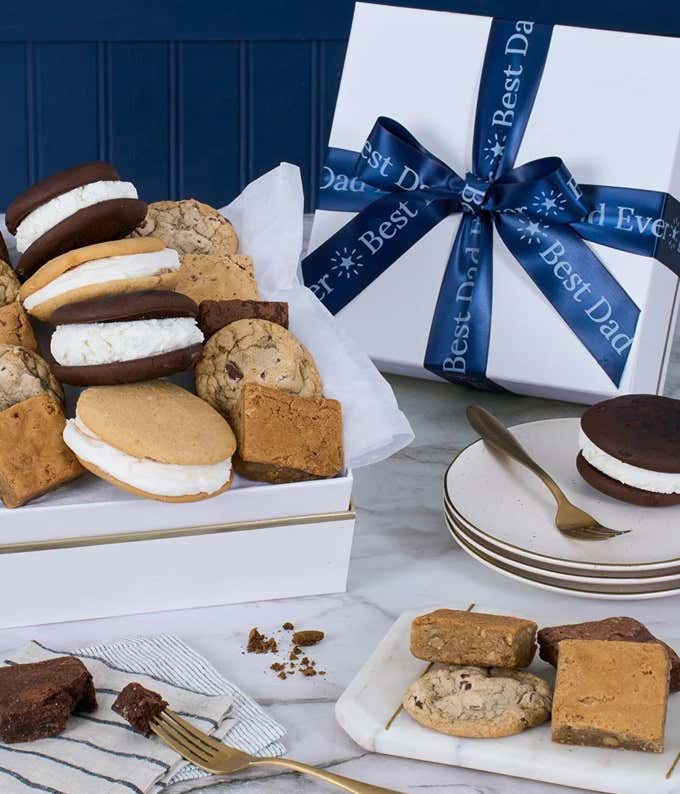 The image features a box labeled 'Best Dad Ever' filled with an assortment of cookies and brownies. The treats are arranged on a marble and wood cutting board, with additional cookies on a small plate nearby.