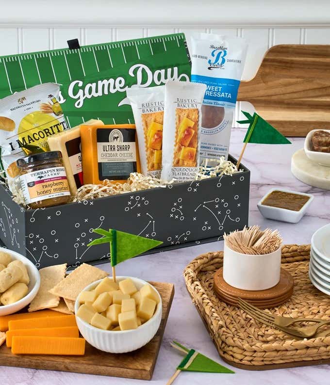 The image shows a snack box with cheddar cheese blocks, crackers, raspberry honey mustard, and other snacks, all packed in a football-themed container. A small football field cutout and foam finger prop add a festive touch.
