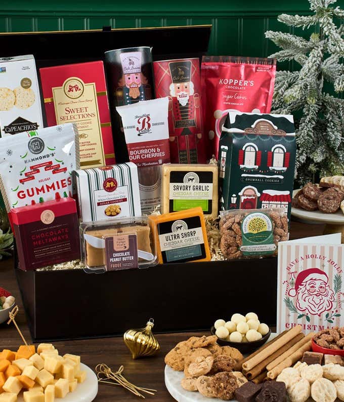 Holiday gift basket filled with assorted cheeses, crackers, cookies, chocolate-covered nuts, fudge, holiday gummies, savory spreads, and candies, all arranged in a black gift box surrounded by holiday decor and ornaments.