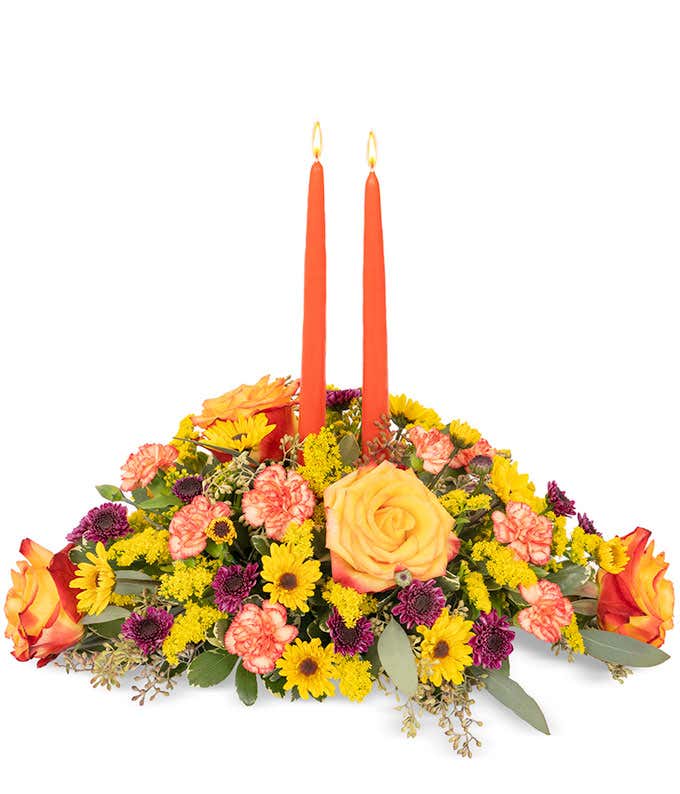 Floral centerpiece full of roses, daisies and carnations in fall colors of orange, purple, and yellow with two tapered orange candles.