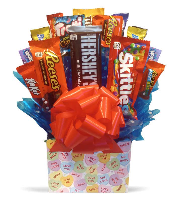 A candy bouquet featuring an assortment of candy bars, including Reese's, Kit Kat, Hershey's Milk Chocolate, Skittles, and Nerds, arranged on blue tissue paper in a colorful container with conversation heart candy designs. A large red bow on the front.