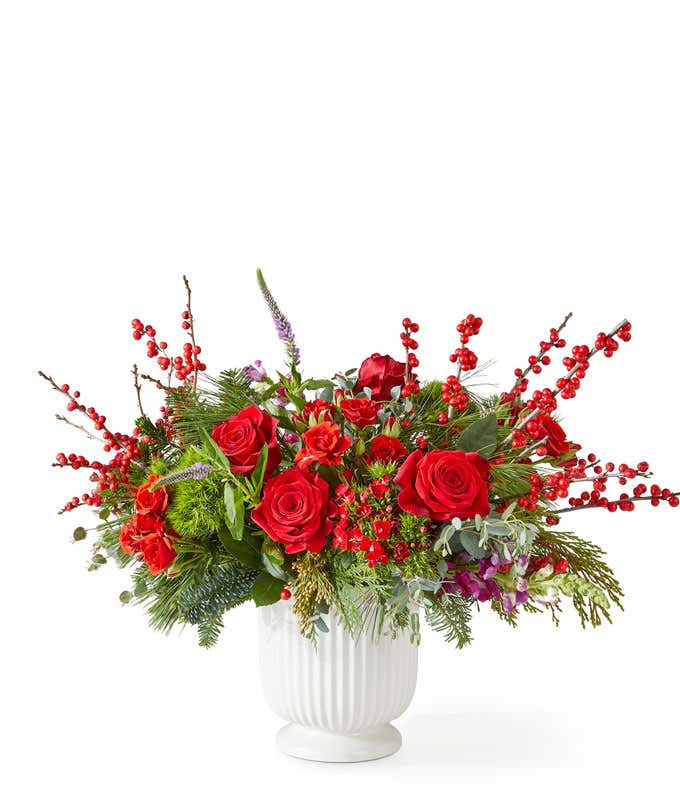 Festive holiday bouquet with red roses, red spray roses, burgundy snapdragons, and burgundy sweet william arranged with pine greenery and red berry accents in a white ribbed ceramic vase.