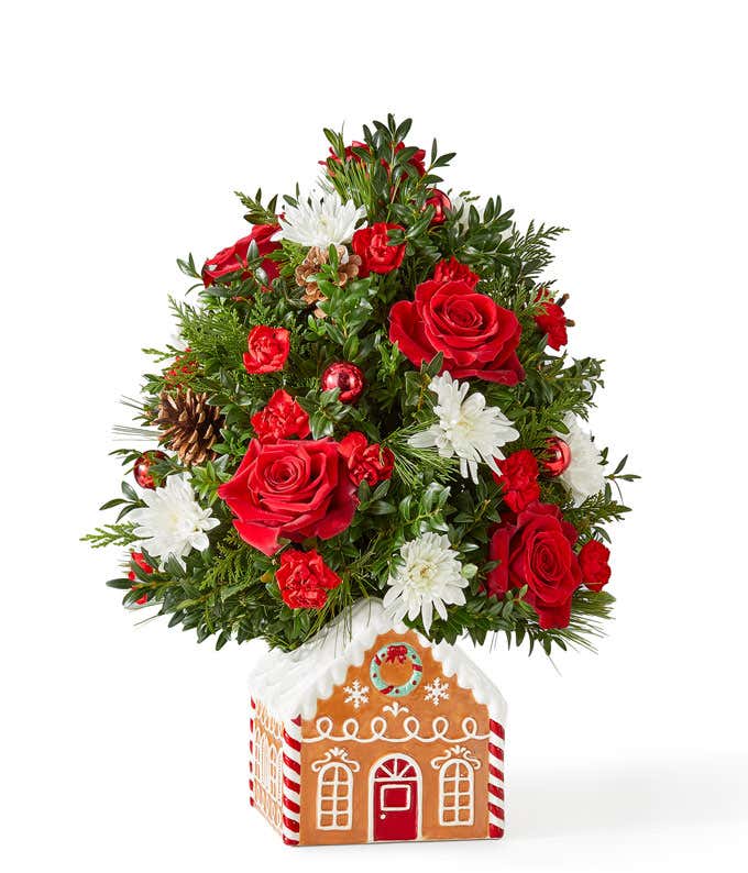 Mini Christmas tree arrangement featuring red roses, miniature red carnations, and white cushion poms accented with festive greenery, pinecones, and red ornaments. Displayed in a charming gingerbread house container.