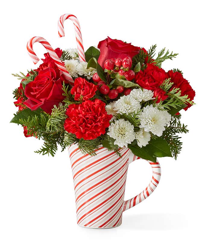 Holiday bouquet featuring red roses, red carnations, miniature red carnations, and white button poms accented with seasonal greenery and two candy canes. Arranged in a white mug with red candy cane stripes.