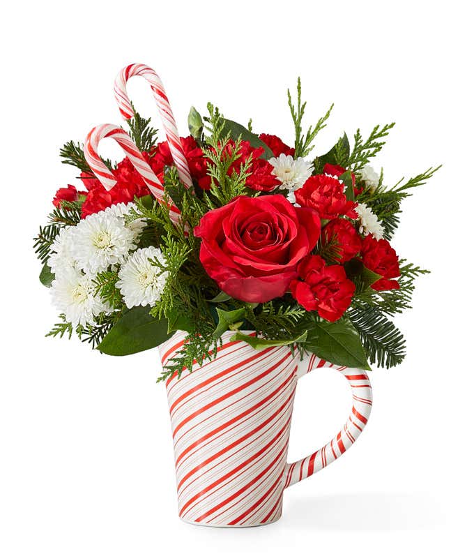 Holiday bouquet featuring red roses, red carnations, miniature red carnations, and white button poms accented with seasonal greenery and two candy canes. Arranged in a white mug with red candy cane stripes.