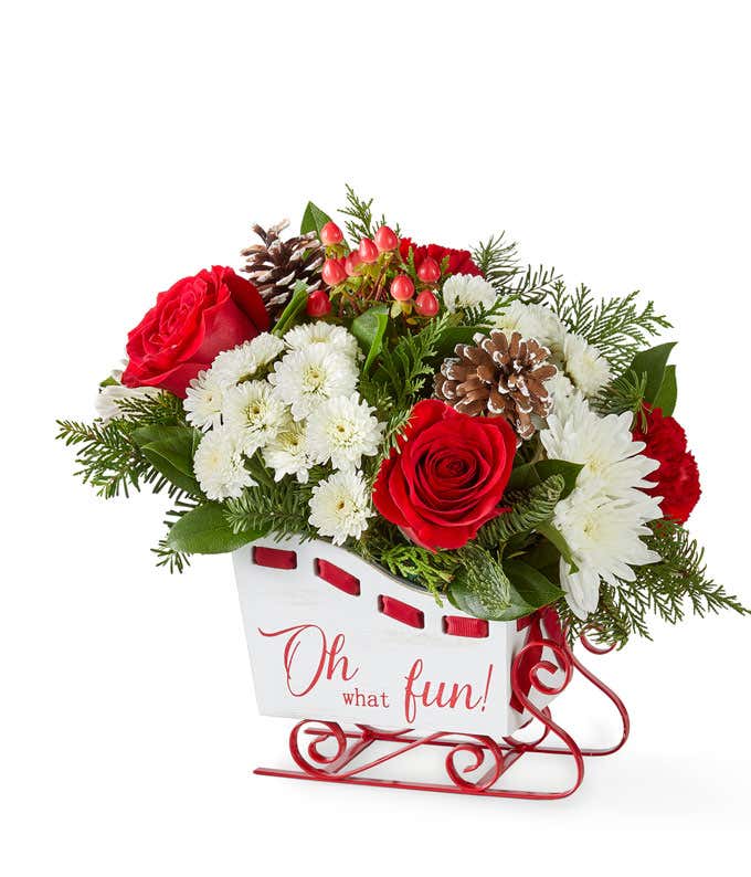 Festive holiday bouquet featuring red roses, red carnations, white button poms, and white cushion poms accented with lush seasonal greenery. Arranged in a cheerful container, this classic red and white floral display captures the joyful spirit of the seas