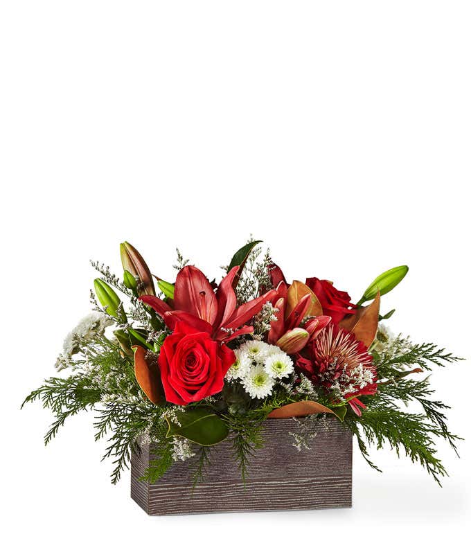 Holiday floral arrangement featuring red roses, lilies, white poms, and greenery artistically arranged in a rustic wooden container.