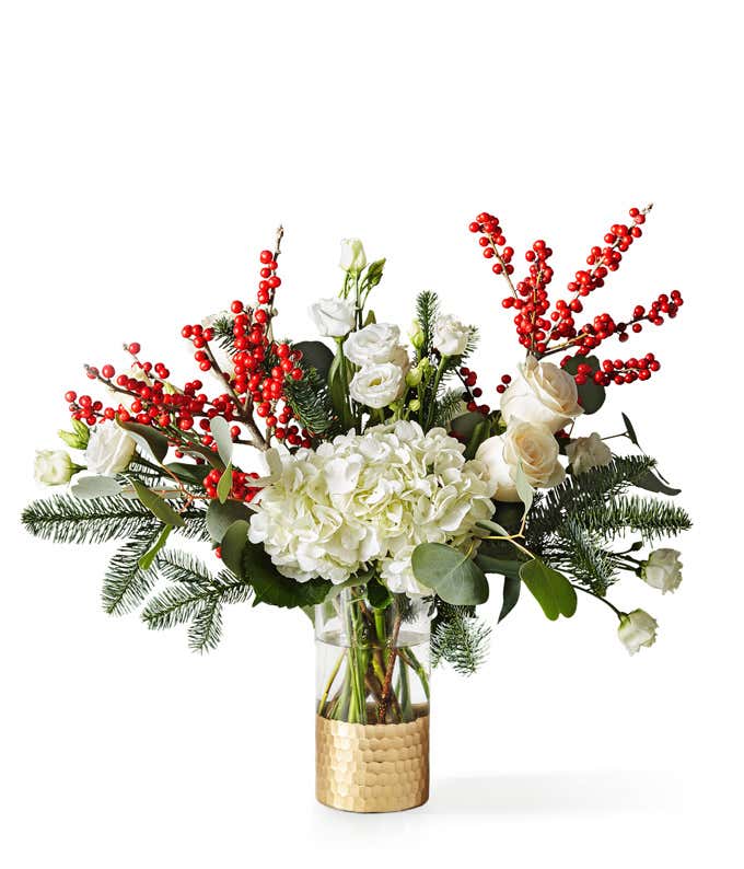Festive winter bouquet featuring white hydrangeas, roses, and lisianthus accented with bright red berry branches, lush greenery, and pine sprigs arranged in a clear vase with a hammered gold base.