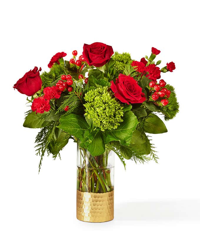 Holiday bouquet featuring red roses, red carnations, green hydrangea, red berries, and seasonal greenery arranged in a clear glass vase with a gold metallic base.