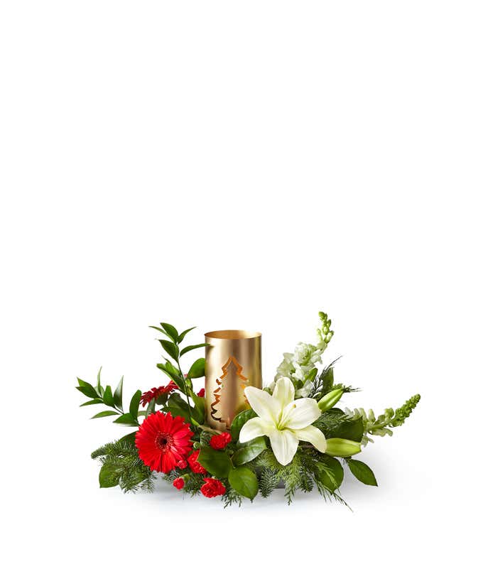 Holiday centerpiece featuring white lilies, red gerbera daisies, red miniature carnations, snapdragons, and greenery arranged around a gold lantern with a Christmas tree cutout design.
