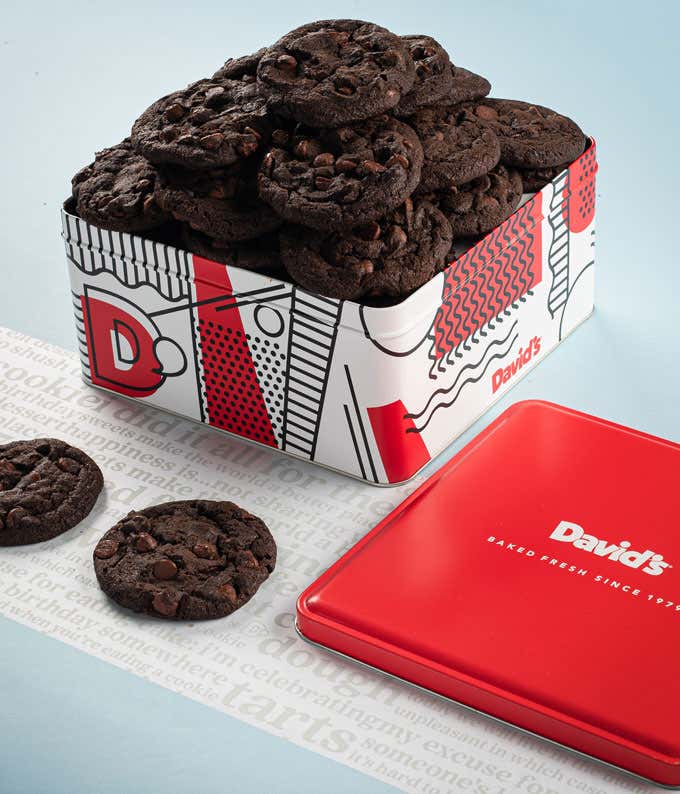 A decorative tin filled with David's double chocolate cookies. The tin features a red, white, and black design with the David's logo. A few cookies and the red lid are placed beside the tin.