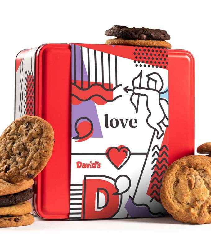 A red tin from David's with a love-themed design, featuring illustrations of a cupid and hearts. The tin is surrounded by assorted cookies, including chocolate chip and double chocolate, with a few cookies stacked on top and beside the tin.