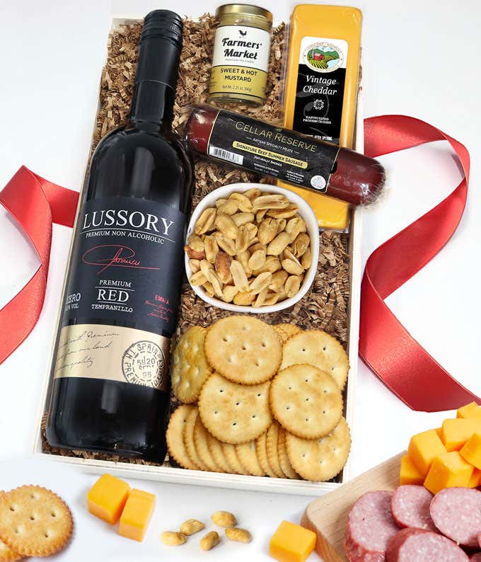 Gift box of cheese, sausage, peanuts, and crackers next to a bottle of red wine