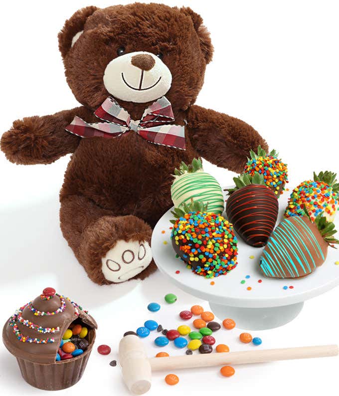 A teddy bear plush next to a breakable chocolate cupcake with a wooden mallet on a wooden serving tray, and 6 chocolate dipped strawberries with drizzles and sprinkles