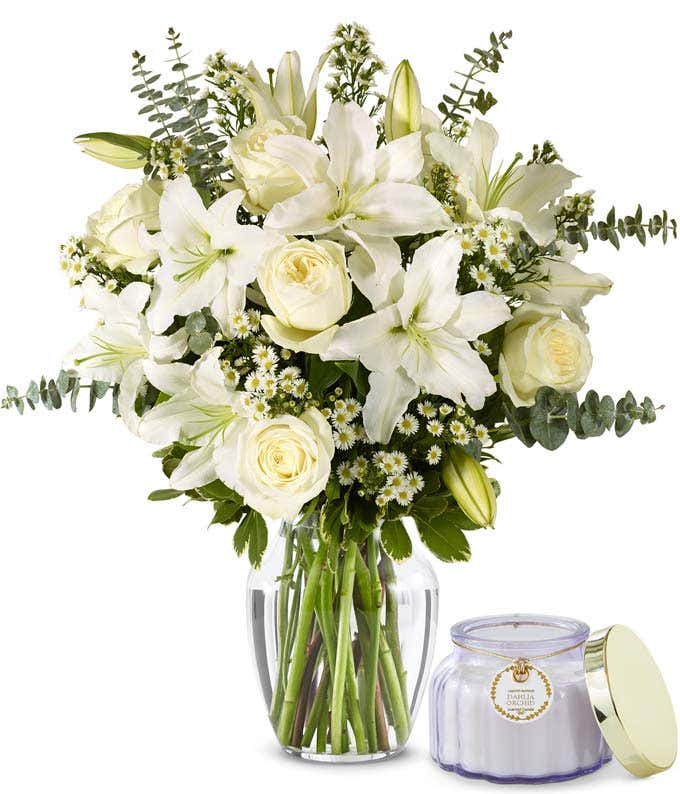 An elegant floral arrangement featuring white roses, white lilies, white asters, and eucalyptus, optionally displayed in a glass vase. Sitting next to a purple candle