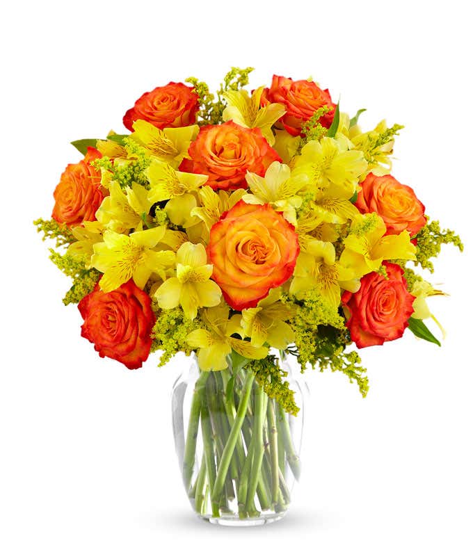 A bouquet of yellow alstroemeria, orange roses with lighter orange centers, yellow solidago in a clear vase on a white background.