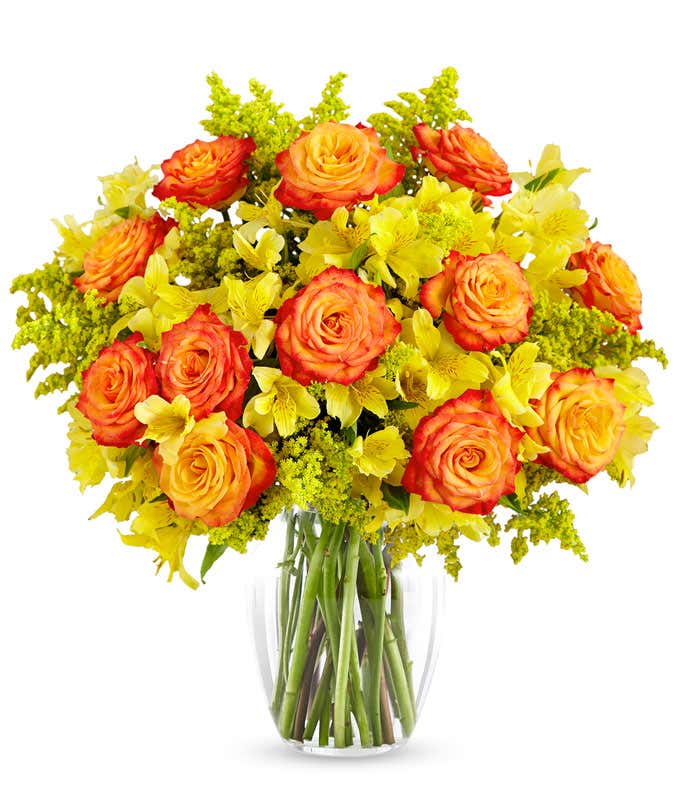 A large bouquet of yellow alstroemeria, orange roses with lighter orange centers, yellow solidago in a clear vase on a white background.