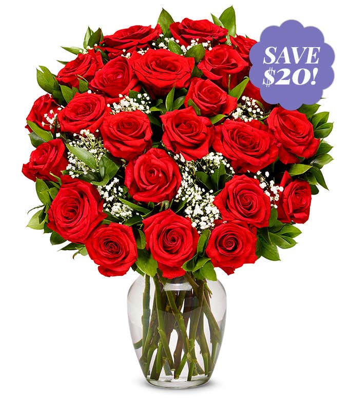 A bouquet of two dozen roses in a glass vase. Between the roses, a few green leaves and bunches of babies breath are visible. 