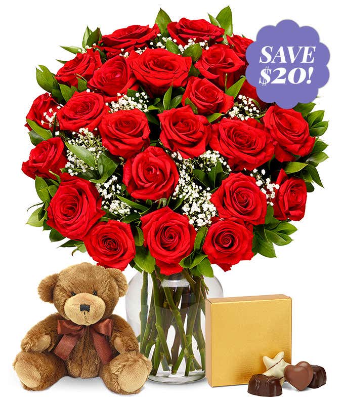 A bouquet of two dozen roses in a glass vase. Between the roses, a few green leaves and bunches of babies breath are visible. Next to the bouquet sits a brown plush teddy bear and a small gold box of chocolates.