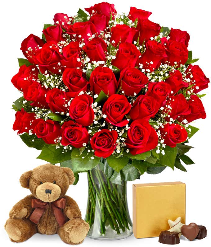 Three dozen vibrant red roses with velvety petals arranged in a clear glass vase with leafy greenery accents. Next to it are a gold square box containing chocolates and a brown teddy bear.