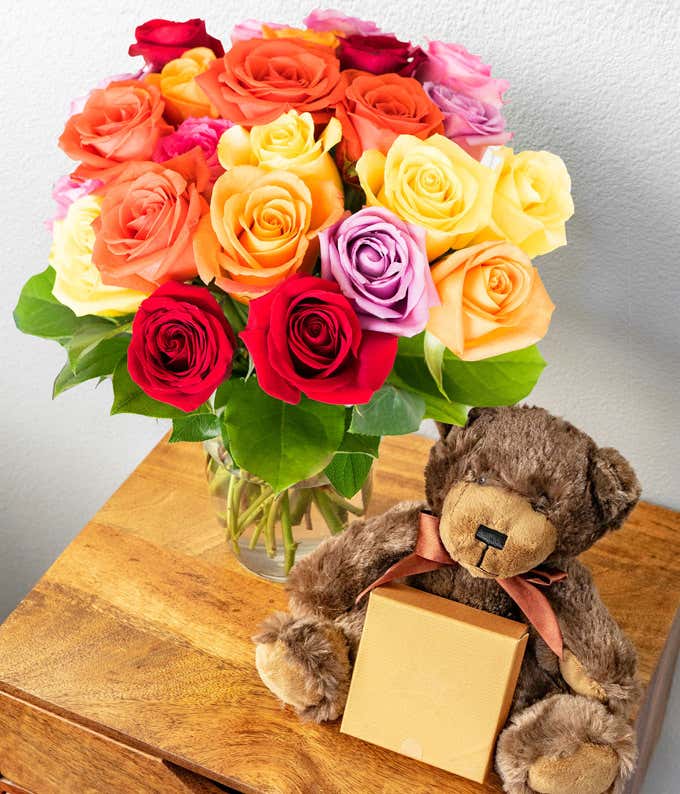 Two Dozen Assorted Rainbow Roses with Chocolates & Teddy Bear in a Clear Glass Vase