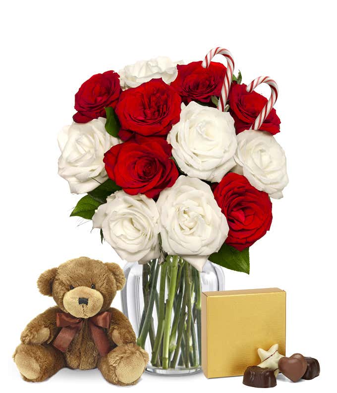 A clear glass vase filled with red and white roses with candy canes, displayed beside a small brown teddy bear and a gold box of assorted chocolates with pieces shown in front.