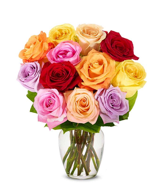 A bouquet of multicolor roses in shades of red, pink, lavender, orange, yellow, and cream, arranged in a clear glass vase with green leaves.