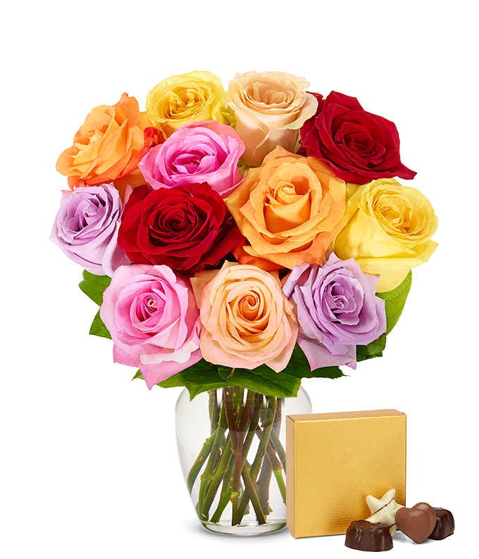A bouquet of one dozen multicolor roses in a clear glass vase, paired with a box of chocolates placed beside the vase.