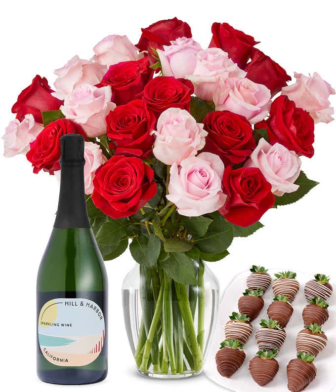 A mixed bouquet of 2 dozen red and blush roses in a clear vase is paired with a bottle of Hill & Harbor Sparkling Wine and 12 chocolate-covered strawberries.