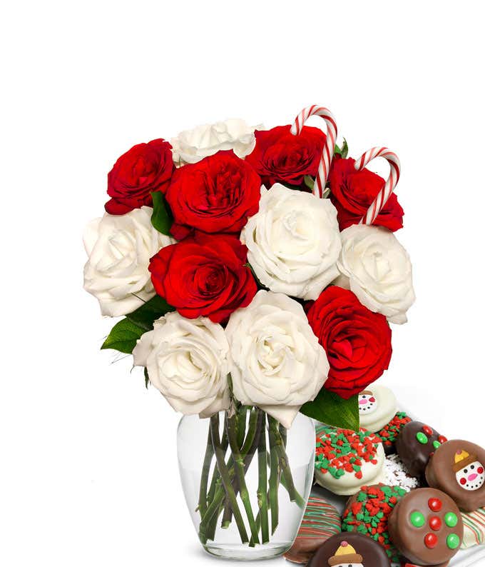 6 red and 6 whtie roses and 2 candy canes popping out of a glass vase with some leaves showing. Next to it is a plate of chocolate cookies with red/green sprinkles, candy, and snowmen.