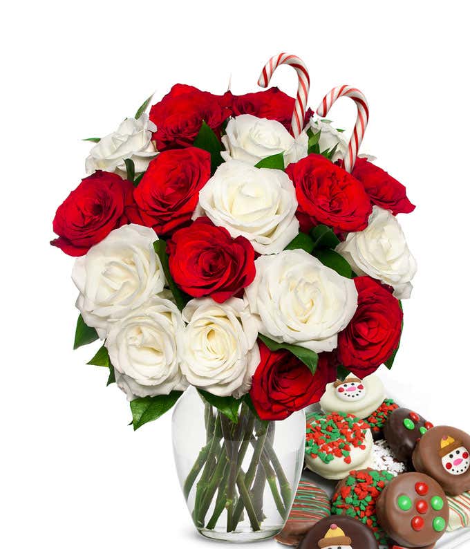 9 red and 9 white roses and 2 candy canes popping out of a glass vase with some leaves showing. Next to it is a plate of chocolate cookies with red/green sprinkles, candy, and snowmen.